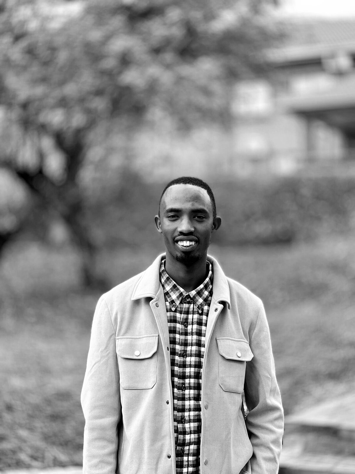 Black-and-white portrait of Caleb Mwenedata smiling outdoors, wearing a light jacket over a checked shirt