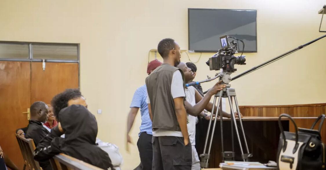 Camera operators filming a courtroom scene with a camera on a tripod while extras sit on wooden benches